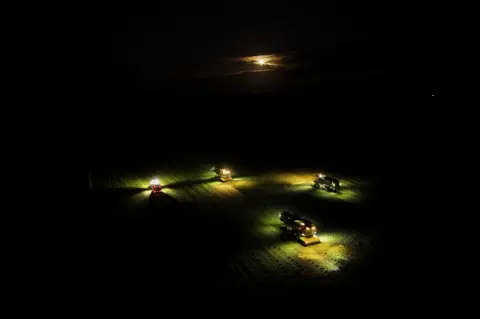 Gerry Priest Aerial image of harvesting machinery with bright lights shining on a field in pitch black darkness with only the moon peaking above the clouds.