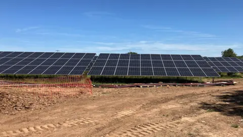 Rows of solar panels at a farm in a field at a farm. The entrance to the field is dry mud with tyre tracks in it.