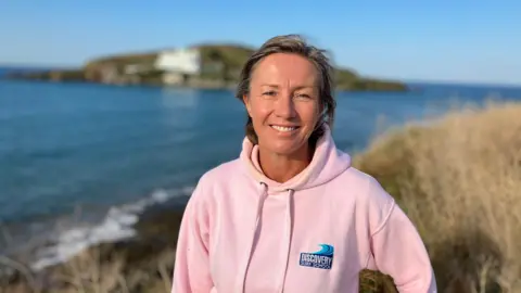 BBC A woman in a pink hoody stands on grassland above a beach. It is sunny and you can see the sea and part of the beach behind her. 