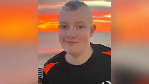 Sarah Waller Aiden standing in front of a vibrant orange sun setting beyond the sea. He wears a black football top and smiles at the camera. He has blue eyes with short cropped lighter hair. 