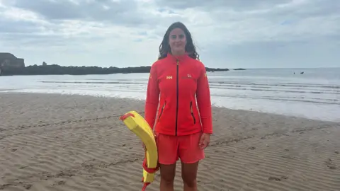 A young female lifeguard wearing a red fleece jacket and red shorts, standing on the beach. The sea is rippling onto the sand behind her.