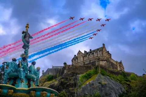 Bob Smart The red arrows flying over Edinburgh castle on an overcast day with the Ross Fountain in Princes St Gardens at the lower left corner. The planes are emitting lines of red, white, and blue smoke as they fly in a V formation.