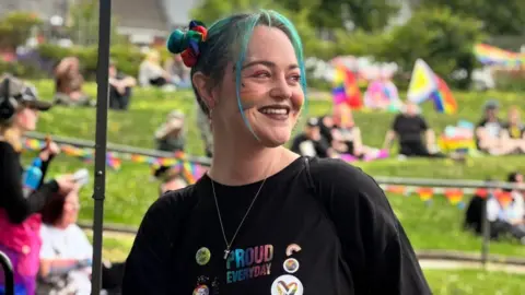 Stephanie Williams Stephanie is wearing her blue hair in pigtails, is in a festival field and is wearing a black t-shirt that says 'proud everyday'. There are people in the background, but they are blurred. She is in focus in the picture. 