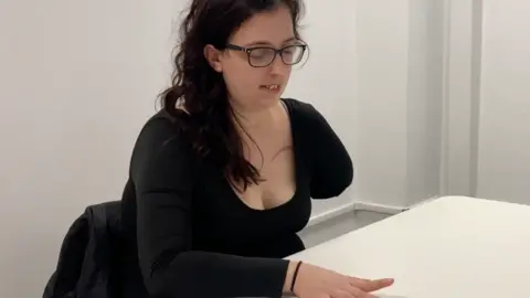 Tamar Makin/Hunter Schone Emily Wheldon, a participant in the study, has long dark hair and is sitting at a white table in a white walled room. She has one hand on the table and is lifting her finger. Her other arm has been amputated. She is wearing a long black top and black framed glasses.