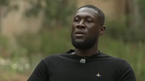 BBC One Show Rapper Stormzy wearing a long-sleeved black T-shirt sits in the grounds of Cambridge University, surrounded by flowers and shrubbery.