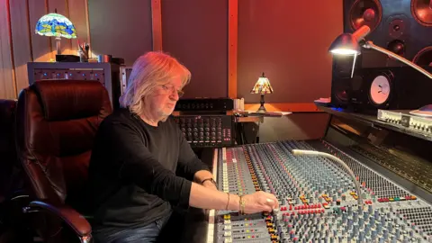 Andy Dransfield A man with shoulder-length blonde hair and glasses sitting in a large black leather chair. He is wearing a black long-sleeved top and jeans. He is leaning over a large sound mixing board and adjusting some of the dials.