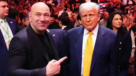 Getty Images Dana White points and smiles at Donald Trump while standing shoulder to shoulder with him at a UFC event. Both are wearing suits and there is a large crowd in the background. 