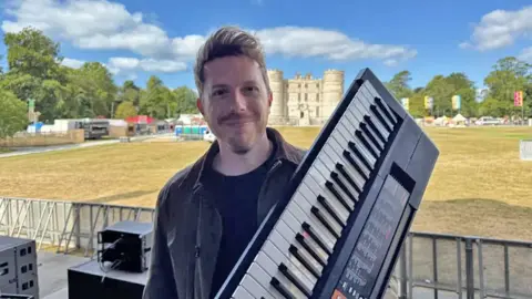 BBC A man with a green coat and blonde hair holding a keyboard in front of a castle and green field.