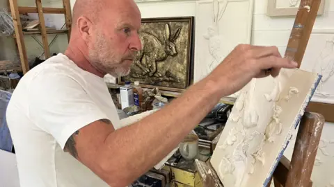 BBC Ian Warren, with a close-cropped greying beard, shaved head and wearing a white t-shirt, works on a piece of art with white plaster on a canvas which is set on an easel. He is in a workshop, with similar artworks of hares on the wall behind him.