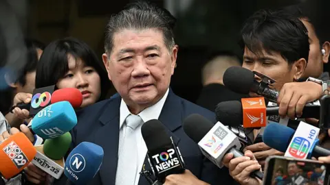 Getty Images Thailand's acting Prime minister Phumtham Wechayachai surroundeed by mics as reporters as ask him questions. He is wearing a balck suit.