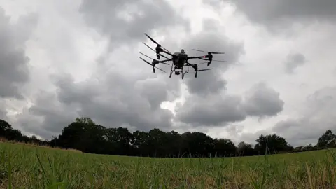 A farming drone flies over a field spraying liquid from four nozzels as it follows a planned route