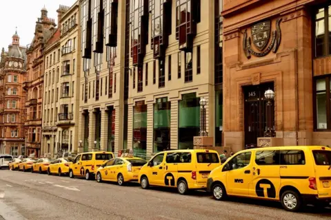 Graham Christie A row of parked yellow taxi's with "NYC" on the side doors, parked outside imposing stone buildings in Glasgow City Centre, styled to look like New York City.