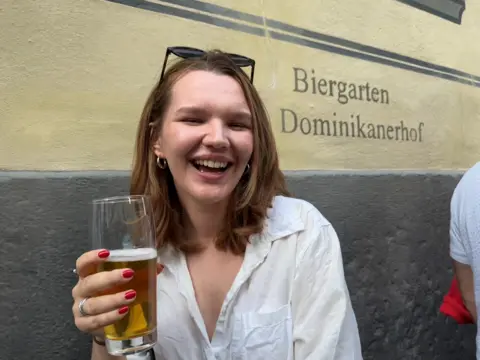 A young lady smiles to the camera holding a half-drunk beer. She has a long auburn bob and red painted nails.
