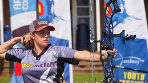 Andrew Scott A woman wearing a had and a sports polo top holding a compound bow up high. Blue archery banners are in the background.