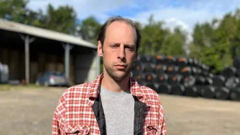 BBC Merlin Lewis , who has brown-hair and is wearing a red checked shirt and grey T-shirt, looks at the camera. There are farm sheds and bales of hay in the background
