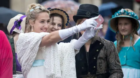 AFP via Getty Images Two young women dressed in outfits from Jane Austen's era smile as they take a selfie during the Jane Austen Festival in Bath. The one most in shot is wearing a white dress with a light blue belt, and has pearl-style earrings. In the background other women in similar outfits are visible