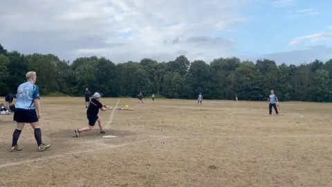 Players from MI6 and the Fuzzy Ducks are mid-game on Wandsworth Common (a dry, grassy field with trees in the background). A female Fuzzy Ducks player wearing a black top, black shorts and black-and-white cap is swinging a bat toward a green-coloured softball.
Behind her stands Gelske Huyer in a light-blue top and dark shorts in the backstop position.
To the right of the image stands MI6's male pitcher in a light-blue coloured top and dark trousers.