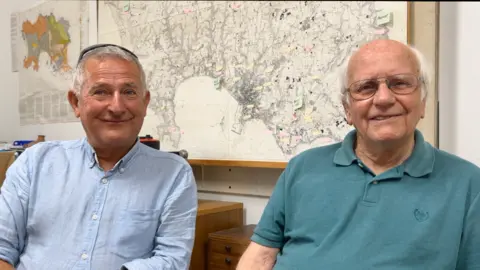 BBC Richard sits on the left in a light blue shirt. Reg is on his right, he is wearing a green polo shirt and has glasses. They both have white hair.
