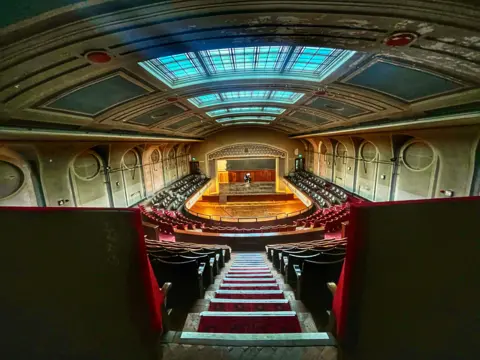 Lara Hughes Old theatre photographed from opposite the stage. up the stairs at the very back. The theatre is empty and has red velvet seats, a slightly curved ceiling with skylight and light green walls.
