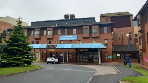 LDRS The exterior of Scarborough Hospital, a brick building of three stories with a blue canopy at the front, a driveway with a white car parked on it and a fir tree to the side.