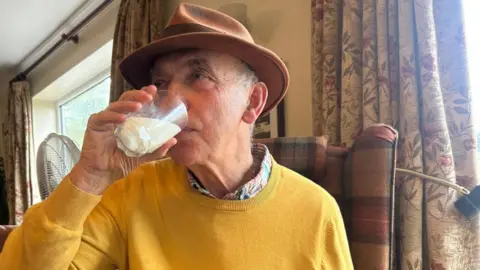 Frank Shellard Frank in an armchair in his living room drinking a glass of mare's milk. He is wearing a yellow jumper and a tan coloured wide brimmed hat.