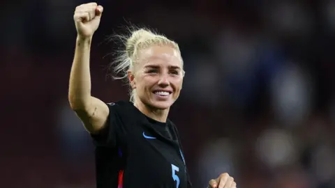 Reuters Alex Greenwood has blonde hair tied back in a ponytail. She has one arm raised in the air and is smiling at the crowd