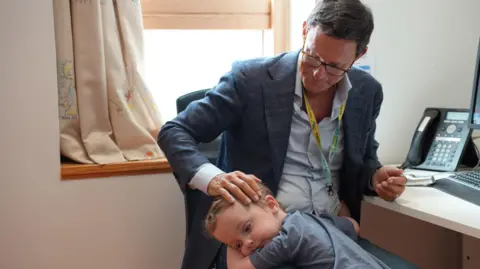 PA Media Conor, now three, from Bolton, hugging his surgeon Conor Mallucci in his office. The surgeon apts his head as he sits at his desk with a phone and keyboard on it.