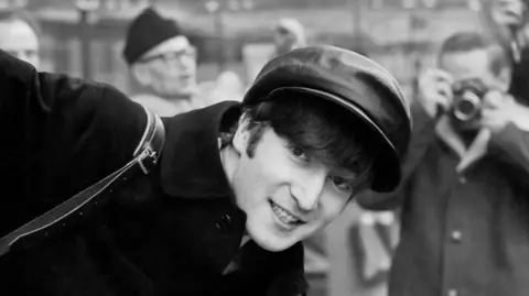 Paul McCartney, Courtesy Gagosian Titled: John on the Champs-Élysées, Paris, 15 January 1964. John wears a bakerboy-style hat, and a dark coat. He leans into the camera frame and smiles. A photographer is seen, blurred in the background.