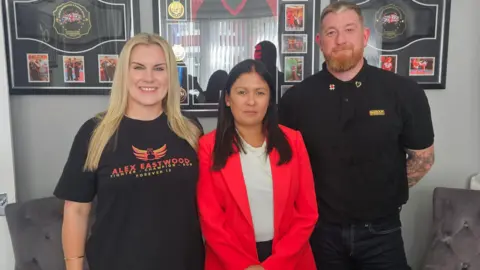 Leigh Day Image shows Nikita Eastwood, left, standing with Lisa Nandy, centre, and Mrs Eastwood's husband Ste in the living room of their home in Liverpool. Mr and Mrs Eastwood are wearing black T-shirts - Mrs Eastwood's bearing her son Alex's name - while Lisa Nandy is wearing a red jacket. There is boxing memorabilia on the wall.
