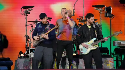PA Media Chris Martin of Coldplay is singing on a stage, with bassist Guy Berryman playing on his right, guitarist Jonny Buckland on his left, and drummer Will Champion playing in the background. Martin is wearing an orange, blue and purple tye-dye T-shirt, with the others wearing dark-coloured jackets. The screen behind them is lit up in bright orange, pink and green patterns.