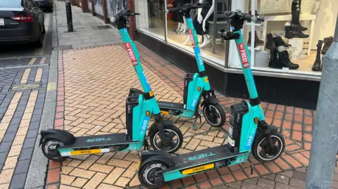Simon Dedman/BBC Turquoise coloured e-scooters - there are three of them - parked on a pavement outside a clothes shop on a paved street.