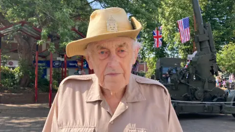 BBC David Shopland looks at the camera with a serious expression. He wears a pale beige military shirt and a distinctive pale yellow hat with a wide rim and a military badge on the front. Behind him a camouflaged green military vehicle can be seen, as well as a Union Jack and the US flag hanging from a tree. 