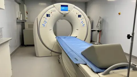 James Paget University Hospitals A CT scanner, which is a large circular machine with a blue display screen at the top. A blue cover is on a bed, as is a wedge-shaped pillow. The walls are white.