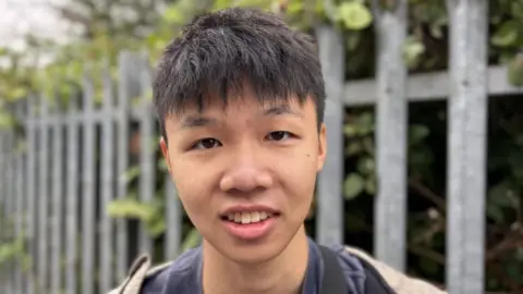 Jasmine Lowe/BBC A man with short black hair with a fringe is smiling into the camera in a head shot. Behind him is a blurred background of a silver fence and trees. He is wearing a navy top and jumper.