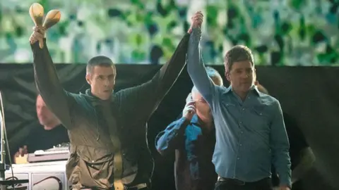 Getty Images Two men raise each others hands. The man on the left is wearing a dark green raincoat and holds two shakers in his right hand. The man on the right wears a blue denim shirt, top button un-done, They both have short brown hair. Behind them are several men who stand in front of a screen displaying green patterns. 