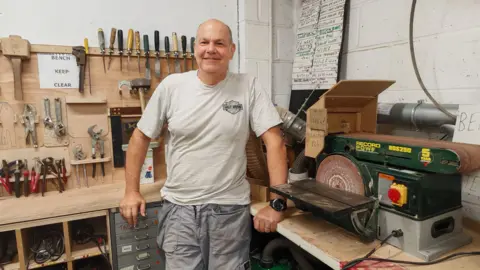 Spelthorne Shed Mike Wallin stands in the Spelthorne Shed. He is wearing a grey t-shirt and grey work trousers. He leans against a work bench, with a circular saw to his left and several racks of woodworking tools to his right. He is a bald middle-aged man and is smiling.