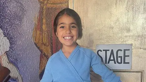 Family handout Alice Aguiar with long brown hair tied back wearing a pale blue dance outfit standing in a theatre in front of a gold door with the sign stage. She is smiling.