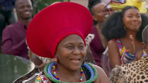 Netflix / Alamy Nandi Nyembe appearing in a film in which she is wearing a bright red traditional hat and beaded necklaces.