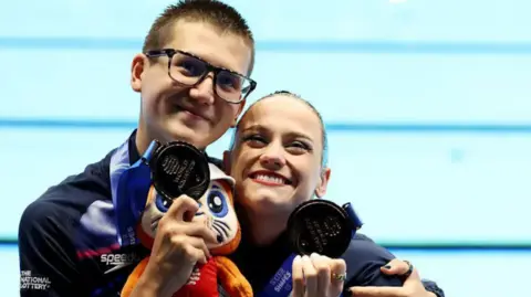 Getty Images Ranjuo Tomblin and Izzy Thorpe stand side by side with their arms around each other in front of a pool. They are smiling and looking up while holding bronze medals up to their faces.
