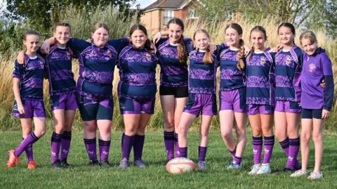 John Gallagher Ten girls, all under the age of 12, stand in a line on a grass pitch in their striped purple jerseys with their arms around each other's shoulders. They smile at the camera, with a rugby ball on the grass in the middle of them.