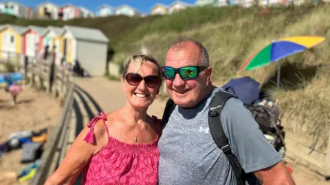 A couple stand on a raised walkway, next to a beach. The sun is shining and they are standing in front of colourful beach huts 