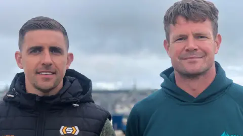 BBC Darryl McDermott is wearing a black gilet with hood with a white and yellow coloured logo. He has short brown hair and stubble. 
Darryl McDermott, who is standing beside Dar