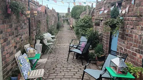 Liz Voce-Jules The cleared alleyway with number of handiong baskets and planters. Seven garden chairs and three small garden tables chairs are lined along the walls which feature planters and hanging baskets. Bunting is hung across the alley.