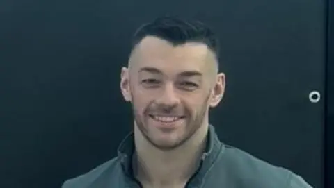 Josh Irwin Josh Irwin is standing behind a black backdrop and is smiling. 
He has short black hair and is wearing a grey jacket and black T-shirt. 