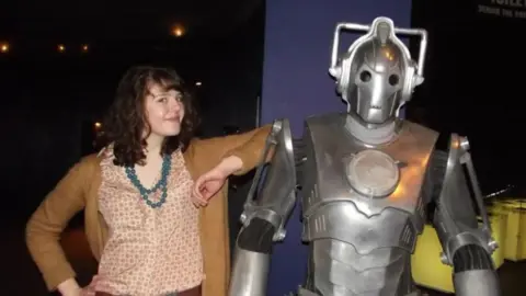 Beth Searby A teenager with curly shoulder-length brown hair smiles, in a brown cardigan and patterned shirt and blue necklace, with her elbow resting on a Cyberman