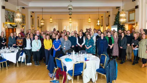 NHS Cornwall and Isles of Scilly Several people stood in a large function room. There are tables in front of some of them. They are all smiling.