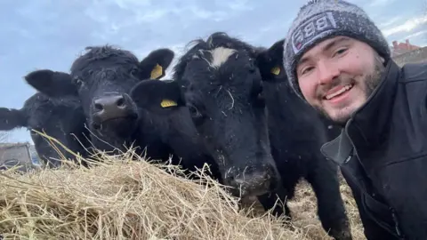 Harry Churchill Harry Churchill in a selfie, on the right hand side of the picture and smiling as he leans into a black cow which is looking at the camera and leaning on another black cow that is also looking at the camera. There is straw in the foreground of the picture and you can see the blue sky behind. 