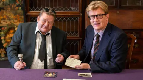 Bearnes Hampton & Littlewood The image shows Dean Buffini wearing  in a dark suit with black tie, which is loosened and dark glasses on his head. He is sitting besides Brian Goodison-Blanks wearing a navy suit and glasses who is holding a gun licence issue to Ronnie Kray in 1964. 
There is a purple cloth on the table in front of them.  Mr Buffini is holding a ring and there is a colour photo featuring his step-father Charlie Kray in front of him and a black and white photo to his right.