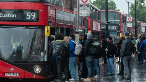 Getty Images A queue of people stand in a rainy London street waiting to board a bus. There are bus stops and other buses visible in the background.