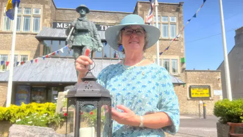 A woman in a blue sun hat and blue patterned top holds a lantern with a candle inside. Behind her is a bronze statue of a soldier. She is in an urban setting, with a large stone building and large flagpoles behind her.
